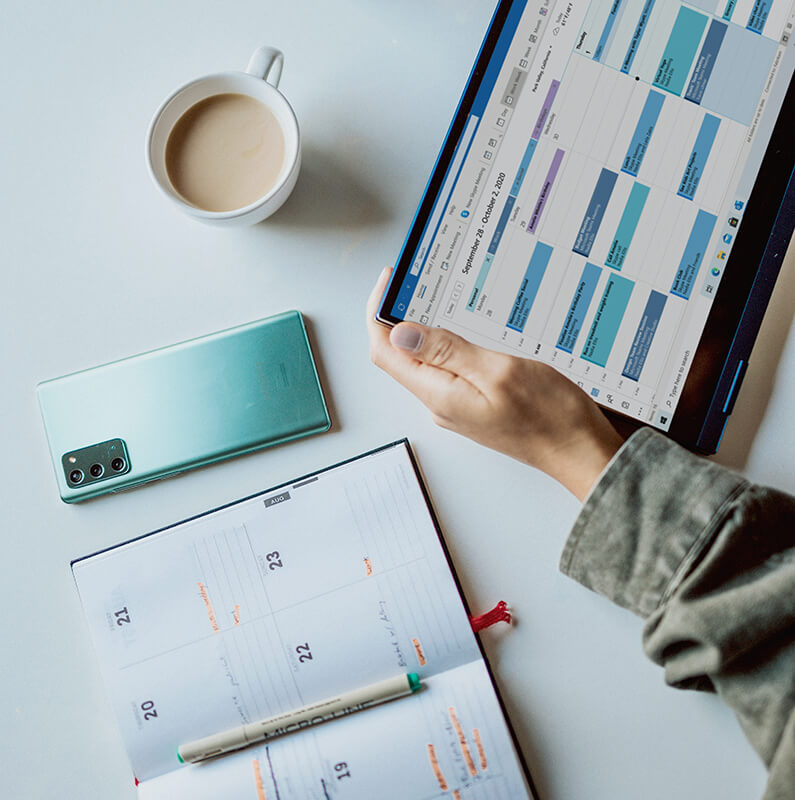 Someone holding a tablet with a schedule with a diary, phone and cup of tea around them